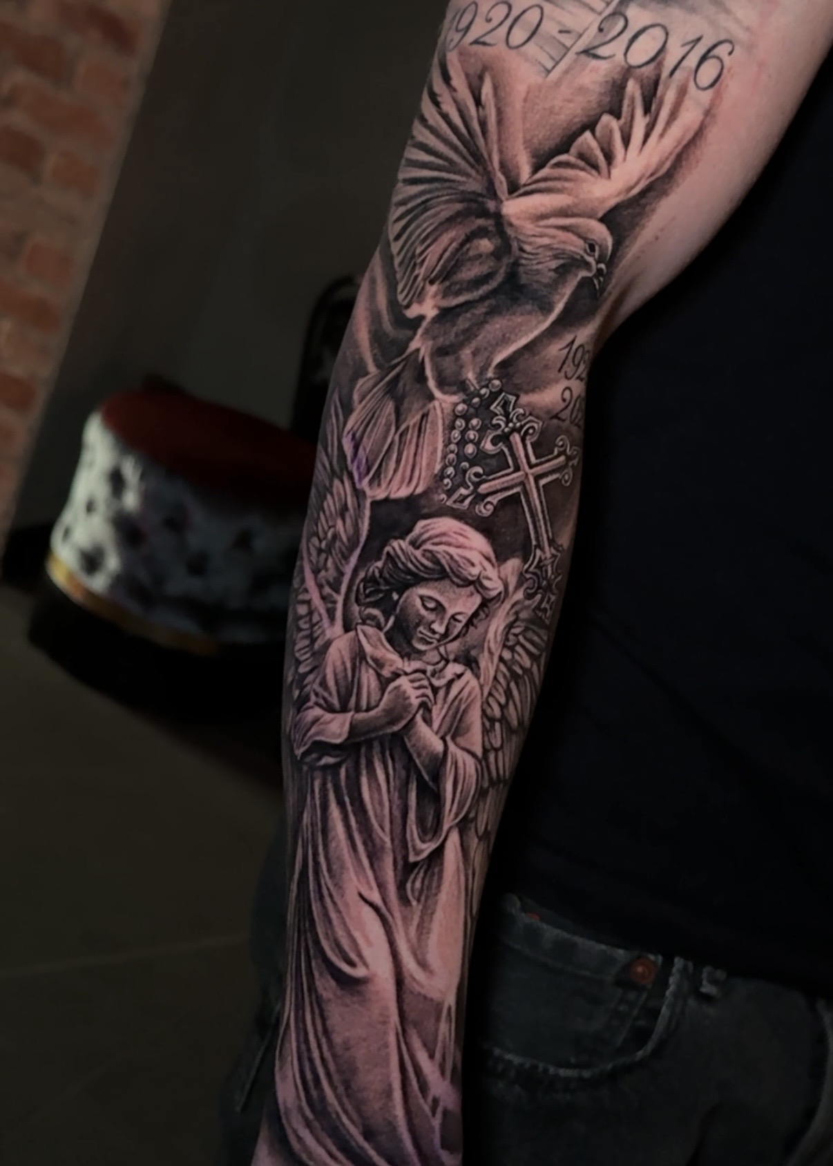 Black and grey-tatuering av ängel och duva med minnesdatum på underarmen